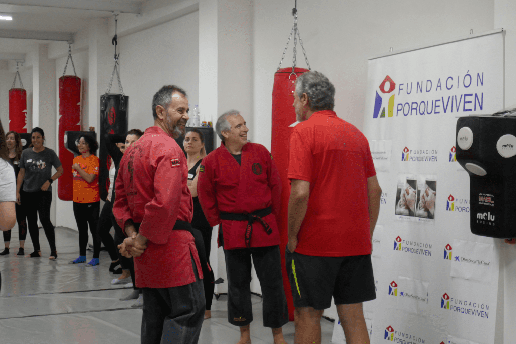 Tres hombres con uniformes de artes marciales conversan frente a un cartel Fundación porqueViven, en un gimnasio donde varias mujeres observan, destacando el compromiso