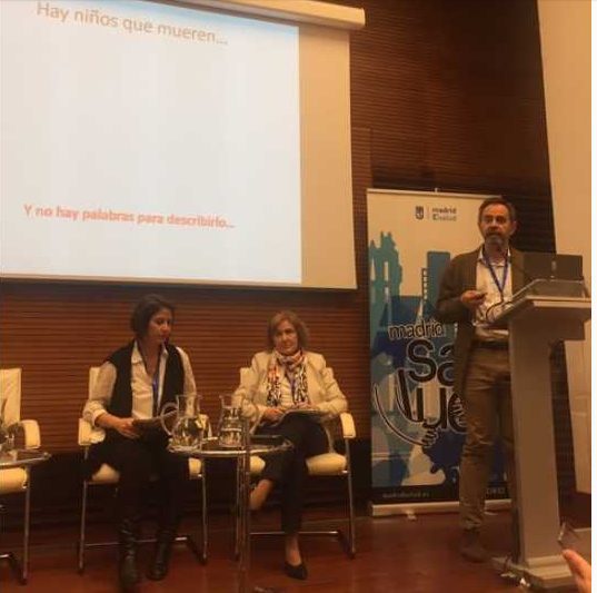 En un panel sobre , organizado por , dos mujeres escuchan mientras un hombre interviene desde el atril; detrás, una proyección con texto en español y el cartel de Madrid Salud