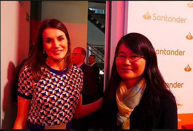 Dos mujeres, una con vestido estampado y otra con gafas y bufanda, en un evento . De fondo, logotipo Santander y asistentes profesionales bajo luz rojiza