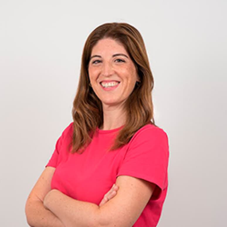 Mujer sonriente de cabello castaño largo, brazos cruzados y camiseta rosa , dedicada