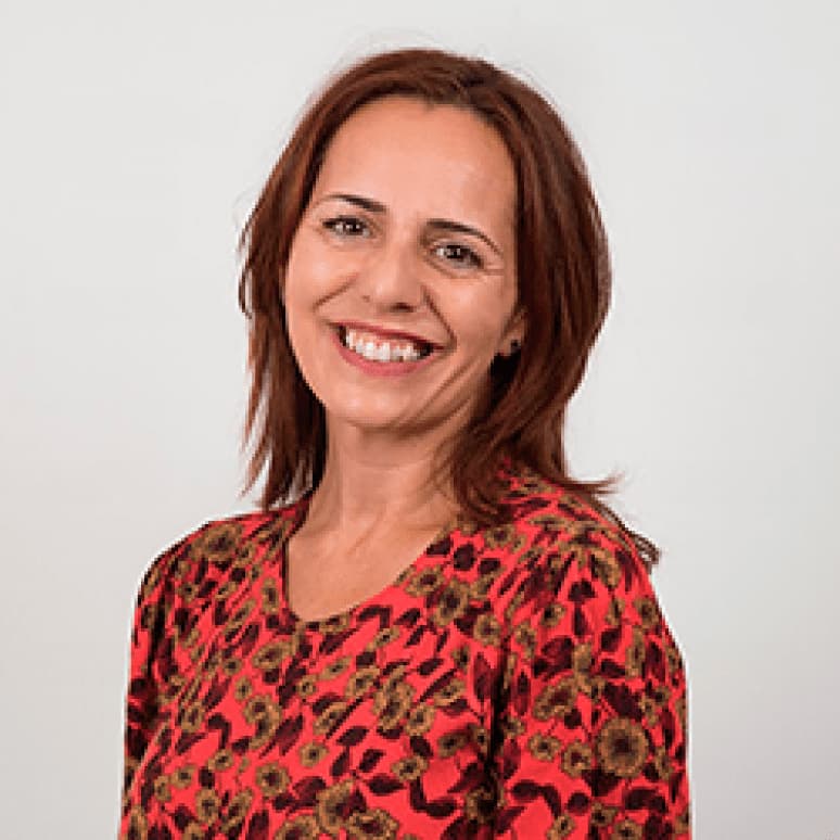 Mujer de cabello castaño sonríe con calidez, lleva blusa floral roja. Fondo claro refuerza su vínculo