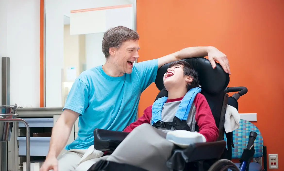En una sala luminosa de paredes naranjas y blancas, un adulto acompaña con una sonrisa a un niño en silla de ruedas; apoya el acompañamiento familiar