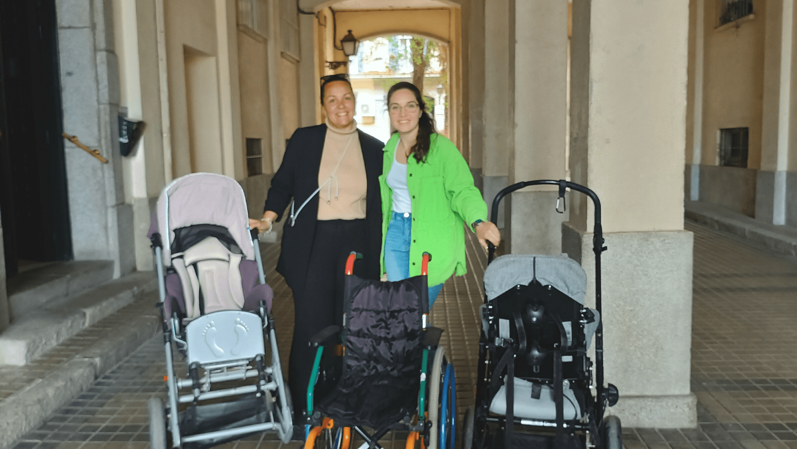En un pasillo exterior, dos cuidadoras Fundación porqueViven sonríen junto a varias sillas de ruedas pediátricas