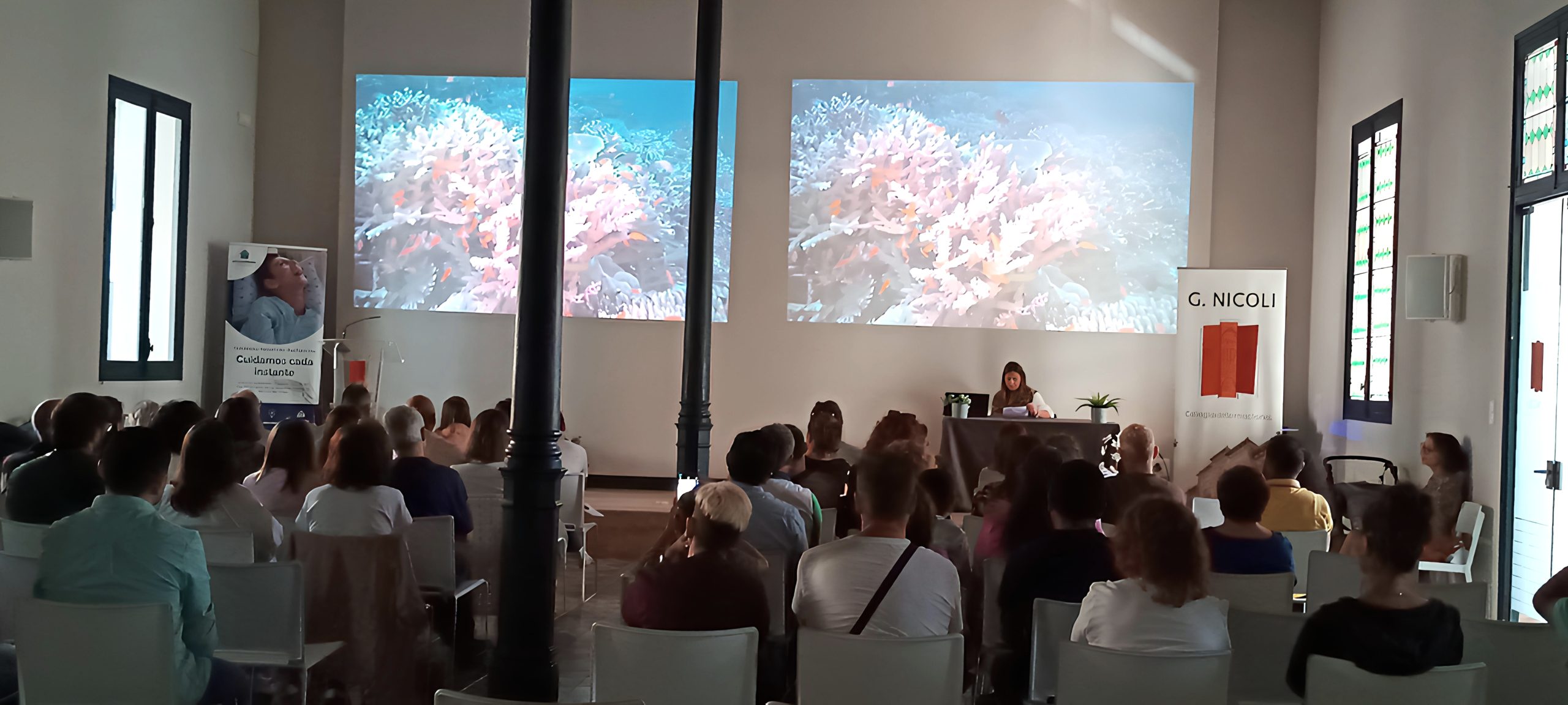 En una sala luminosa, una representante Fundación porqueViven habla a familias tras la pérdida de sus hijos; pantallas y pancartas rodean el espacio