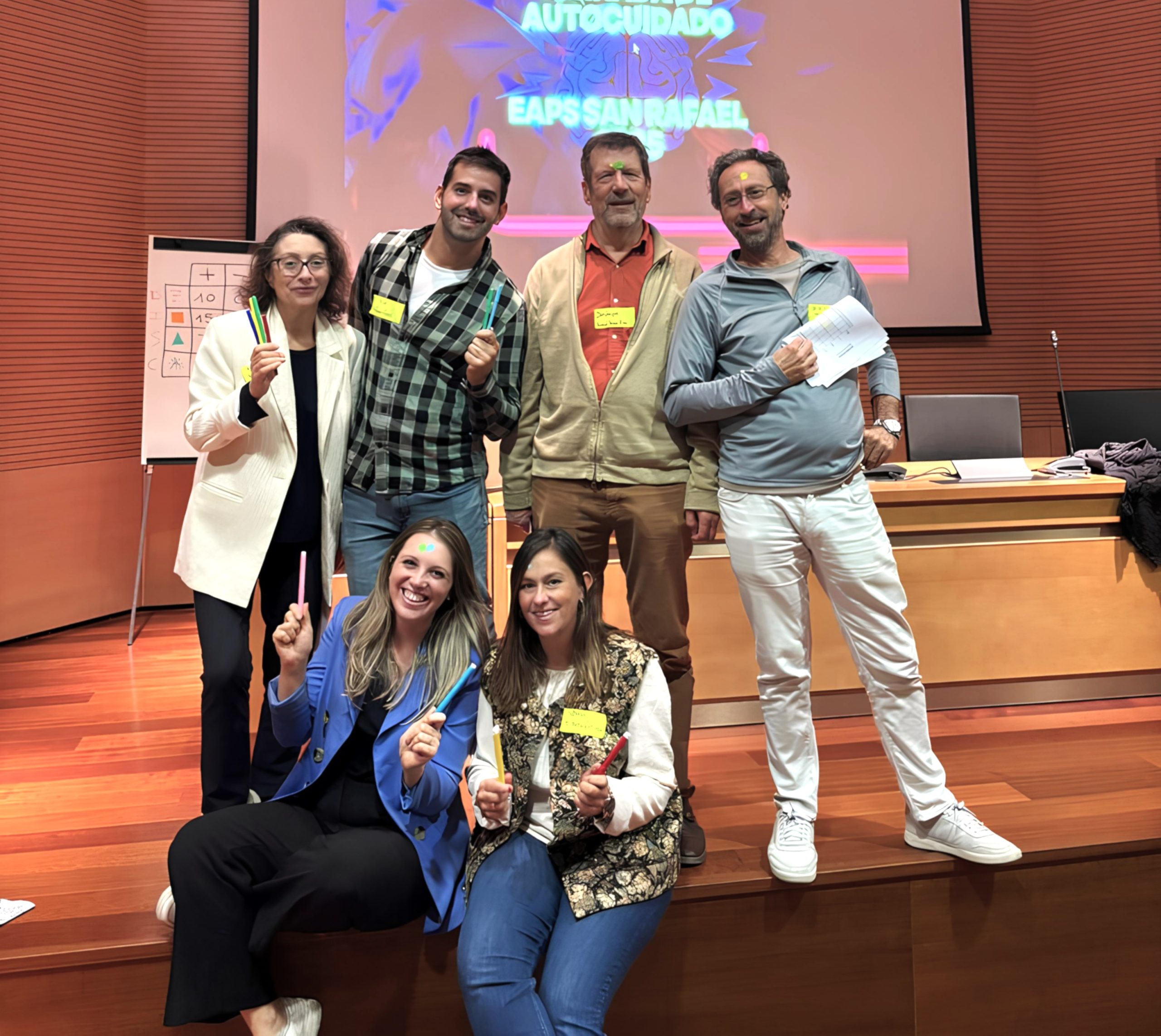 En un evento de , seis profesionales de la posan en el escenario, mostrando bolígrafos de autocuidado junto a una mesa con material y una pantalla informativa