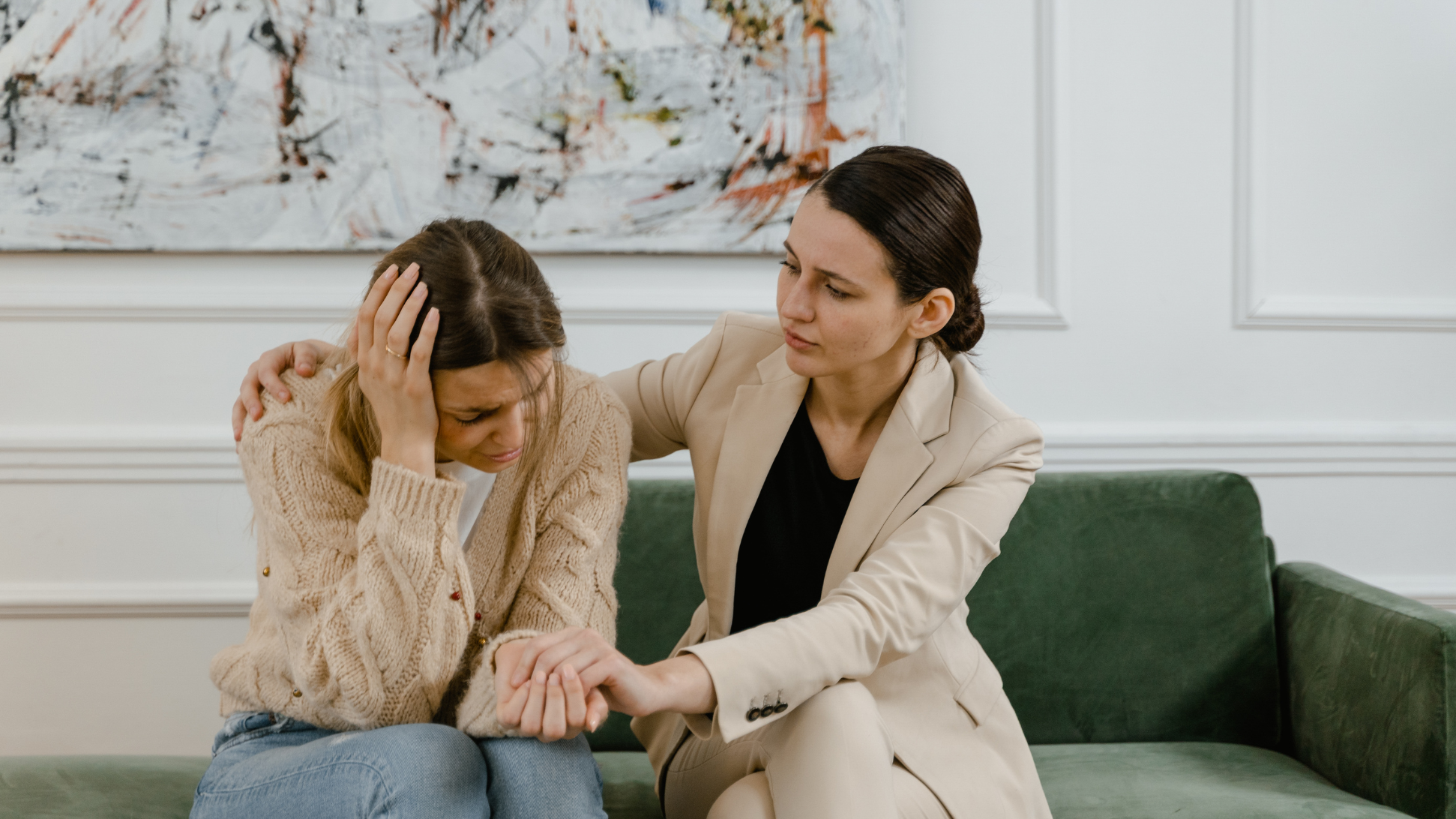 Dos mujeres sentadas en un sofá verde. Una parece angustiada, sujetándose la cabeza, mientras la otra -vestida de traje y con experiencia en voluntariado y vida personal- la consuela cogiéndole suavemente del brazo y ofreciéndole su preocupación.